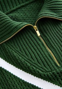 Zelený pletený svetr s žebrovanou strukturou, bílým vodorovným pruhem a částečně rozepnutým zlatým zipem u límce.