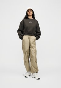 Νεαρή γυναίκα που στέκεται με τα χέρια στις τσέπες, φοράει σκούρο φούτερ New Balance, μπεζ παντελόνι cargo, λευκό καπέλο και λευκά αθλητικά παπούτσια.