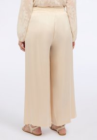 Ljusbeige vida byxor i lätt tyg, med elastisk midja och slät yta, bärs tillsammans med enkla sandaler.