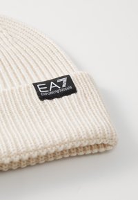 Szydełkowy beanie w kolorze kremowym, z teksturą w prążki i czarną metką z logo "EA7 Emporio Armani" wyszytym z przodu.