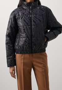 Black puffer jacket featuring a glossy finish with repeated logo pattern, high collar, and elasticized hem, paired with beige trousers.
