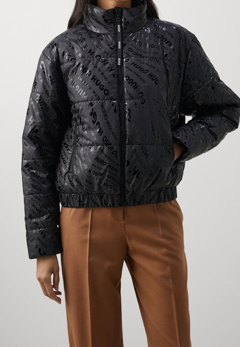 Black puffer jacket featuring a glossy finish with repeated logo pattern, high collar, and elasticized hem, paired with beige trousers.
