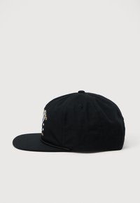 Cappellino da baseball nero con visiera piatta e design ricamato in oro e bianco parzialmente visibile sul lato anteriore sinistro.