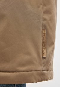 Καφέ ύφασμα που δείχνει μια κοντινή εικόνα του τε�λειώματος ενός σακακιού, με μια ταμπέλα σε ταμπά χρώμα που φέρει χαραγμένο το λογότυπο "Marc O'Polo." Η υφή φαίνεται λεία και ελαφρώς γυαλιστερή.