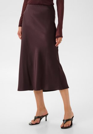 Femme portant une jupe midi marron foncé, un top transparent à manches longues et des sandales à talons noires avec les orteils découverts sur un fond uni.