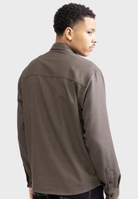 Brown checkered shirt with a classic collar, long sleeves, and buttoned cuffs. Fabric has a textured, woven pattern.