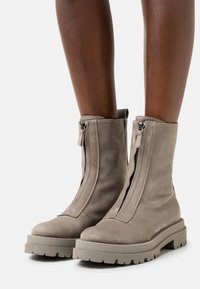 Bottes de cheville en daim taupe avec des semelles en caoutchouc épaisses et des fermetures éclair à l'avant, portées par une personne à la peau foncée.