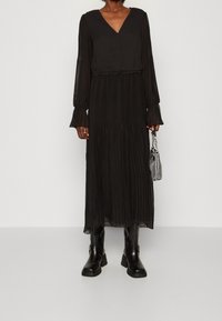 Svart plisserad maxiklänning med långa ärmar, V-ringning och detalj med insamlad midja. Bärs med svarta knähöga läderstövlar och en liten väska.