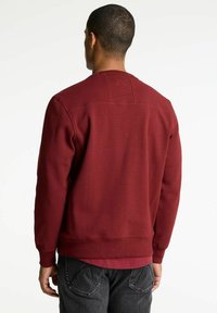 Uomo con capelli corti che indossa un sweatshirt bordeaux e jeans neri, in piedi di spalle contro uno sfondo chiaro e uniforme.