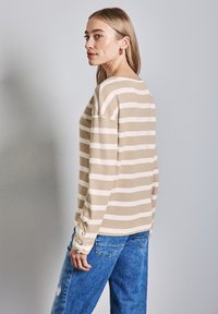 Beige- und weiß gestreiftes Langarmoberteil mit Rundhalsausschnitt, strukturiertem Stoff und Knopfdetails an den Ärmeln. Kombiniert mit blauen Jeans.