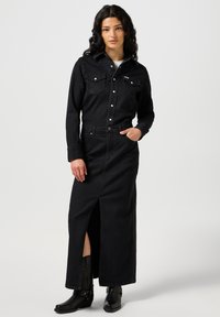 Czarna dżinsowa koszula zapinana na guziki z dwiema kieszeniami na piersiach, w połączeniu z czarną dżinsową spódnicą o wysokim stanie z rozcięciem z przodu, noszoną z czarnymi botkami.