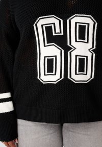 Zwart gebreid sweater met witte nummer 68 op de voorkant, witte strepen op de manchetten, gedragen met lichtgrijze broek door een persoon.