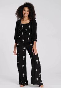 Mono negro con patrón de palmeras blancas; cuerpo fruncido, mangas largas abullonadas, pantalones anchos. Lleva sandalias de tacón abierto.