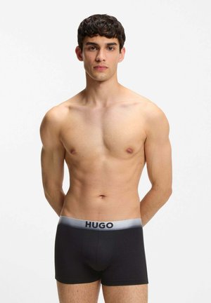 Young man standing straight wearing black boxer briefs with gray waistband labeled "HUGO" against a plain white background.
