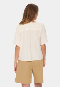Blusa corta a maniche corte in crema con tessuto texture, dettaglio arricciato sul retro e scollo rotondo morbido, abbinata a pantaloni corti marrone chiaro.