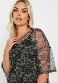 Top in pizzo nero con maniche trasparenti, motivo floreale e scollatura ampia. La texture morbida e la forma fluente conferiscono un aspetto elegante.