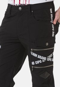 Schwarze Cargo-Hosen mit mehreren Reißverschlusstaschen, Akzenten aus weißem Branding-Tape und einer glatten Textur. Verfügt über einen klassischen Knopfverschluss und Seitennähte.