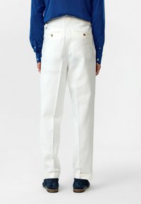 Witte linnen broek met een relaxte pasvorm, voorzien van twee achterzakken en zichtbare stiksels, gecombineerd met marineblauwe suède schoenen.