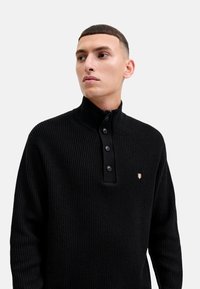 Schwarzer gerippter Strickpullover mit hohem Kragen, ausgestattet mit einer halben Knopfleiste und einem kleinen Emblem auf der Brust.