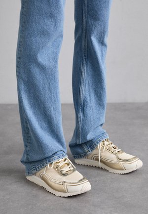 Sneakers beige con accenti dorati metallici, pannelli in rete e suola in gomma testurizzata, abbinati a jeans azzurri lievemente svasati.