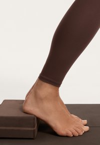 Rjave legice, prikazane od meč navzdol, počivajo na rjavi joga kocki. Noga je gola z gladko, svetlo kožno teksturo.