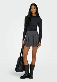 Kvinne som har på seg en svart rynket langermet topp, grå plisserte miniskjørt, svarte støvler til midten av leggen, holder en stor svart totebag, står på hvit bakgrunn.