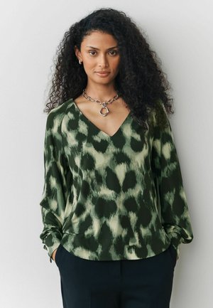 Grön och krämfärgad v-krageblus med leopardmönster, långa ärmar och mjukt tyg. Kombination med mörka byxor och ett silverhalsband.