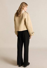Beige cropped jacket with a textured collar, paired with black straight-leg trousers. The jacket features shoulder button accents.