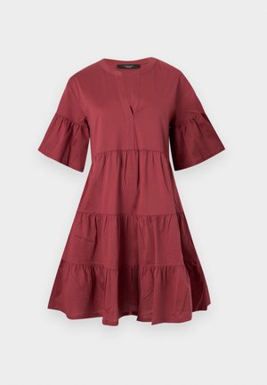 Femme portant une robe rouge à volants, manches courtes, longueur genou, avec un col fendu et des talons noirs pointus, debout sur un fond uni.