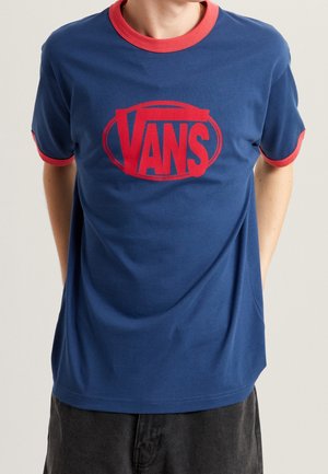Osoba nosząca niebieską koszulkę Vans z czerwonym kołnierzem i obszyciami na rękawach oraz dużym czerwonym logo Vans na środku klatki piersiowej.