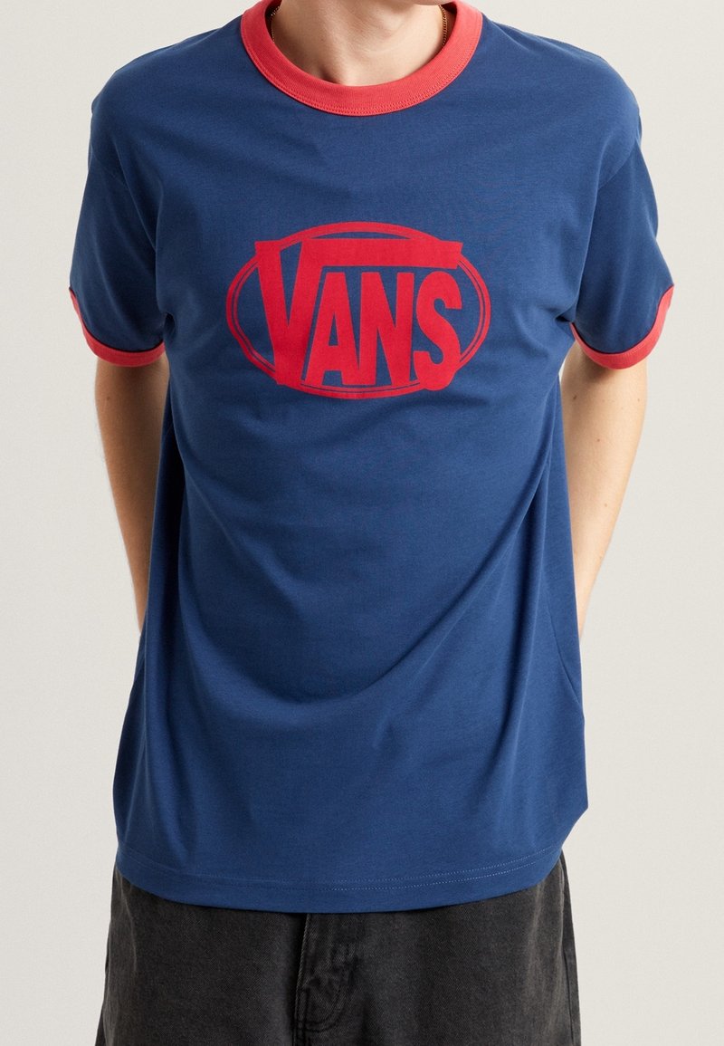 Personne portant un t-shirt bleu Vans avec un col et des bordures de manches rouges, et un grand logo rouge Vans centré sur la poitrine.