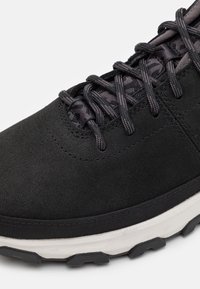 Gros plan d'une basket noire avec une tige en tissu texturé, des lacets noirs, et une épaisse semelle blanche avec des détails en relief noirs.