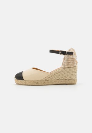 Espadrilla con zeppa, superiore in tessuto beige e punta in gomma nera, dotata di tacco in juta intrecciata e una fascia alla caviglia nera con fibbia.