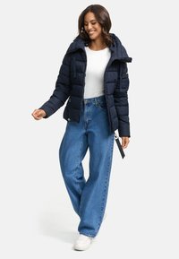 Mariene pufferjack met capuchon, ritssluiting aan de voorkant en elastische manchetten, gecombineerd met hoge taille, wijde blauwe jeans en witte sneakers.