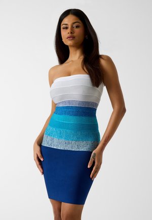 Kobieta o długich ciemnych włosach, ubrana w dopasowaną sukienkę bez ramiączek z poziomymi pasami w kolorze od białego do ciemnoniebieskiego, stojąca na jednolitym tle.