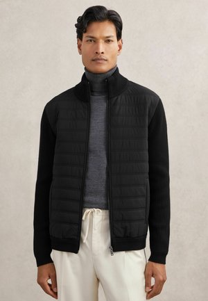 Mand iført en sort quiltet jakke med lynlås over en grå rullekrave og cremefarvede bukser med snøre, stående foran en neutral baggrund.