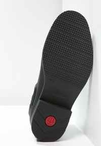 Semelle de chaussure en caoutchouc noir avec une texture antidérapante, portant un logo circulaire rouge et le nom de la marque "HUNTER" en relief. Design lisse et plat.