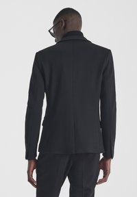 Blazer nero su misura con una texture liscia, spalle strutturate, spacco centrale e bottoni scuri. Presenta un colletto a punta e una vestibilità slim.