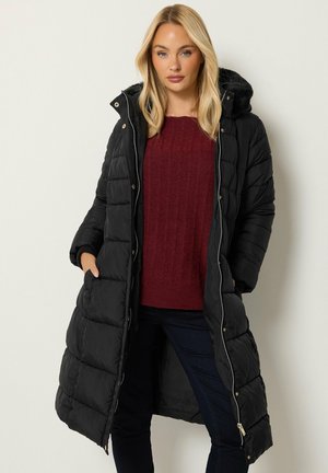 Femme portant un long manteau matelassé noir avec capuche, un pull bordeaux et un pantalon foncé, debout les mains dans les poches du manteau sur un fond clair.