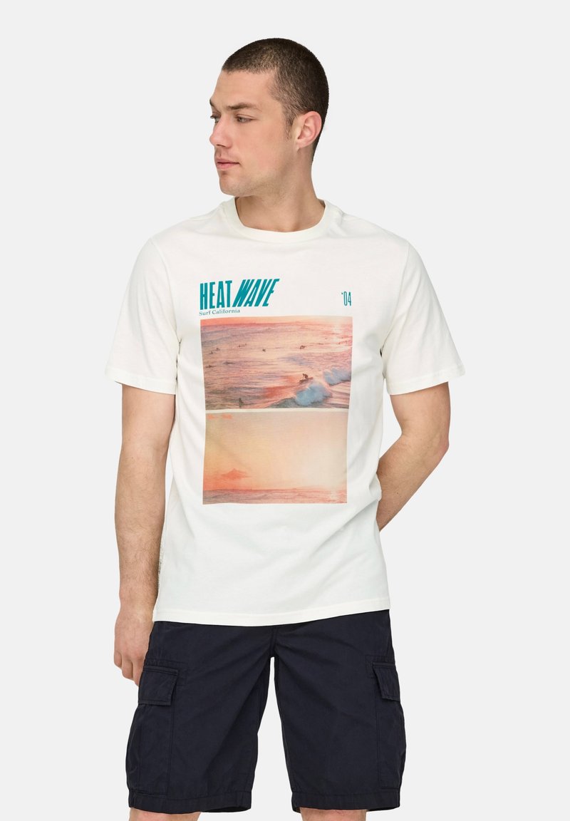 Hombre vistiendo una camiseta blanca con un gráfico de surf y el texto "HEAT WAVE Surf California", combinado con pantalones cortos de carga negros, mirando hacia un lado.