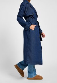 Trench coat de denim azul oscuro con cintura ceñida por un cinturón, cuello con muescas y botones grandes. Presenta bolsillos laterales y un corte relajado.