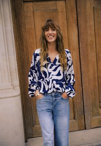 Blau-weiße gemusterte Bluse mit V-Ausschnitt und weiten Ärmeln, kombiniert mit hellblauen, hoch taillierten Jeans. Holz-Hintergrund.