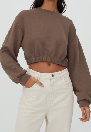 Kvinde iført en brun kort sweatshirt med elastisk kant og cremefarvede højttaljede bukser, stående med den ene hånd i lommen mod en neutral baggrund.