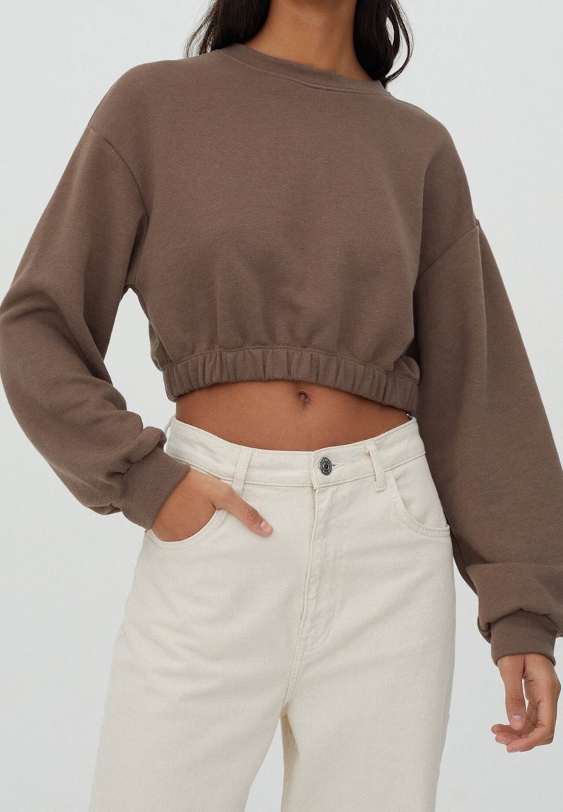Femme portant un sweat-shirt court marron avec un ourlet élastique et un pantalon crème taille haute, debout avec une main dans la poche devant un fond neutre.