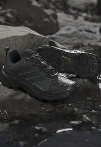 Chaussures de randonnée noires imperméables avec semelles robustes, placées sur un sol rocheux humide, montrant les détails de la bande de roulement et des gouttes d'eau à la surface.
