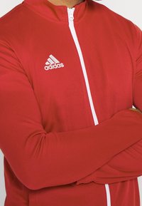 Veste de sport rouge avec un col haut, fermeture éclair blanche et logo Adidas sur la poitrine gauche. Fabriquée en tissu lisse et léger.
