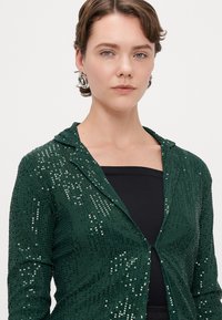 Veste verte à sequins avec capuche, fermeture à boutons sur le devant, et un haut noir en dessous. Texture brillante avec une coupe ajustée.