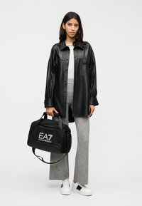 Giacca camicia in pelle nera, pantaloni grigi in maglia, sneaker bianche, borsa nera con logo "EA7 Emporio Armani", design minimalista, silhouette strutturata.