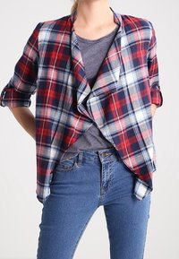 Rutigt skjorta i marinblått, rött och vitt, med en draperad front och korta ärmar, lager på en grå topp och matchat med blå jeans.