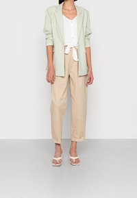 Ljusgrön blazer över en vit blus, knuten vid midjan, i kombination med beige vida byxor och vita sandaler. Släta, skräddarsydda tyger.